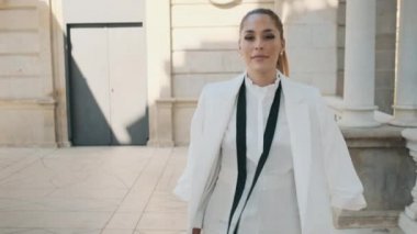 Beyaz takım elbiseli muhteşem zarif kadın kameraya bakıyor. Şehir parkında eski bir mimariyle yürüyor.
