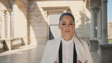 Beyaz takım elbiseli şık bir kadının şehir parkında eski güzel bir mimariyle güvenle yürüdüğünü gösteren bir fotoğraf. Dışarıda poz veren güzel bayan. 