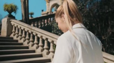 Klasik beyaz takım elbiseli muhteşem genç bir kadının eski şehir parkında merdivenlerden çıkarken ve kamerada beni takip ettiğini gösteren bir fotoğraf.
