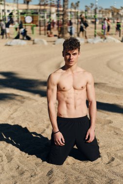 Genç yakışıklı sporcu, çıplak gövdesiyle sıcak yaz günü plajda idmandan sonra kameraya bakıyor. Çekici fitness erkek modeli dışarıda poz veriyor.