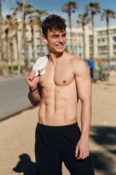 Young positive sporty man with naked torso joyfully resting after workout outdoor. Attractive fit guy happily posing on city beach