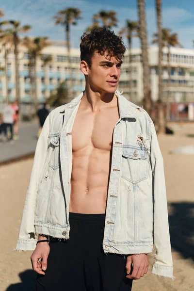 stock image Young confident sporty guy in denim jacket with naked torso posing on camera on city beach