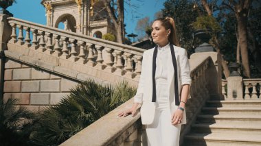 Eski güzel şehir parkında merdivenlerde duran klasik beyaz takım elbiseli muhteşem bir kadın. Zarif model kendine güvenerek dışarıda poz veriyor.