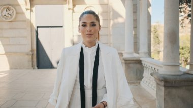 Beyaz takım elbiseli zarif genç bir bayan şehrin parkında eski güzel bir mimariyle güvenle yürüyor. Güzel bayan dışarıda poz veriyor.