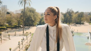 Şehir parkında güzel bir manzarayla poz veren şık beyaz takım elbiseli muhteşem bir kadının portresi.