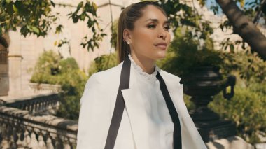 Beyaz ceketli ve tişörtlü muhteşem bir kadın eski güzel şehir parkında güvenle yürüyor. 