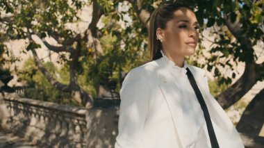 Beyaz ceketli ve tişörtlü zarif bir kadının portresi güvenli bir şekilde eski güzel şehir parkında yürüyor. 