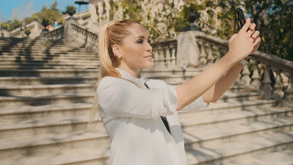 Genç, çekici, zarif bir kadın eski güzel şehir parkında merdivenlerde akıllı telefondan selfie çekiyor. Güzel bayan dışarıda selfie çekiyor.