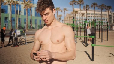 Şehir plajında spor yaparken akıllı telefon kullanan, kablosuz kulaklıklı yakışıklı bir adamın portresi.