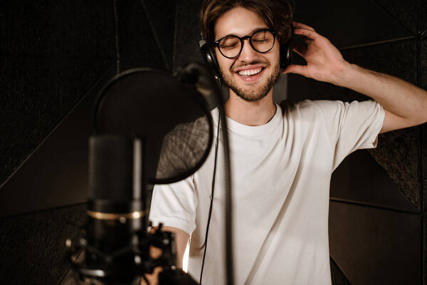 Young attractive male singer in headphones joyfully singing in modern sound studio 