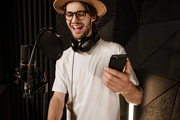 在现代录音室里 年轻迷人的男歌手带着智能手机在感情上歌唱 — 图库照片