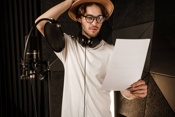 Young thoughtful male singer intently reading text of new song in modern sound recording studio 