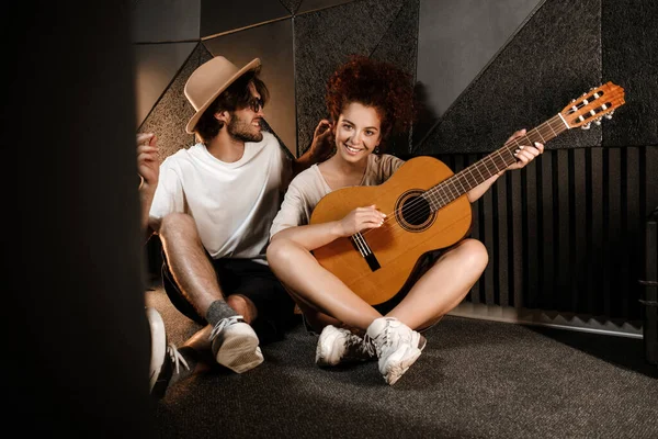 年轻迷人的男男女女坐在现代录音室的地板上快乐地弹着新歌 — 图库照片