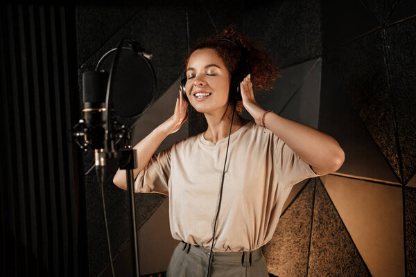 Beautiful smiling singer girl in headphones dreamily recording new song in professional studio