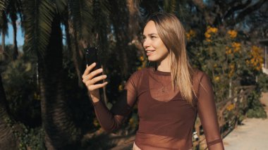 Güzel kız şehir parkında yürürken ailesiyle görüntülü konuşma yapıyor. Açık havada telefon kullanan çekici bir kız.