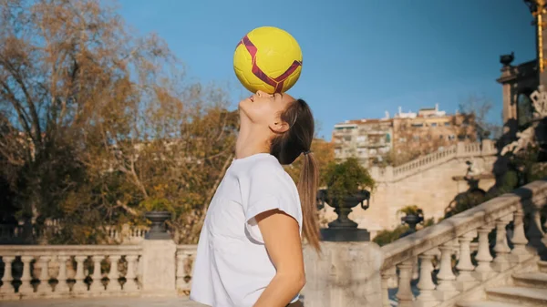 Attractive sporty girl practicing football skills in city park. Beautiful football girl trying to hold ball on head outdoor