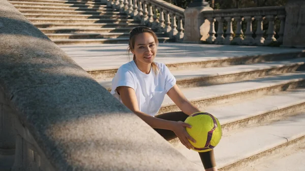 在城市公园的楼梯上 一个漂亮的女运动员在镜头前快乐地看着足球 躺在老楼梯上 — 图库照片