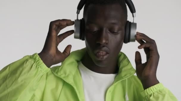 ポートレートのハンサムなスタイリッシュなアフリカ系アメリカ人男で明るいジャケット熱心に聴いて音楽でヘッドフォン以上グレーの背景 — ストック動画