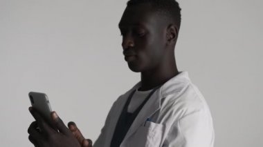Genç ve çekici Afrikalı Amerikalı erkek doktor, gri arka planda telefonda görüntülü konuşma yaparken el sallıyor. Selamlama ifadesi