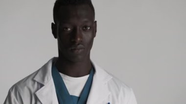 Genç, ciddi, Afro-Amerikalı erkek doktor gri arka plandan kameraya bakıyor. Kızgın ifade