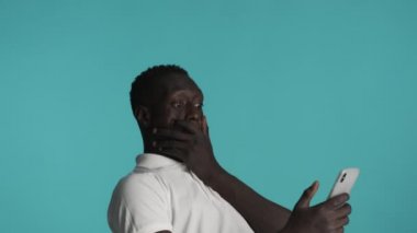 Genç afro-amerikan adam akıllı telefon kullanıyor ve mavi arka planda seviniyor. Mutlu ifade