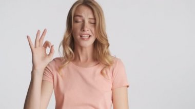 Çekici, neşeli sarışın kız mutlu bir şekilde göz kırpıyor ve gri arka planda kameraya iyi bir jest yapıyor. İfade etrafında dolaşmak