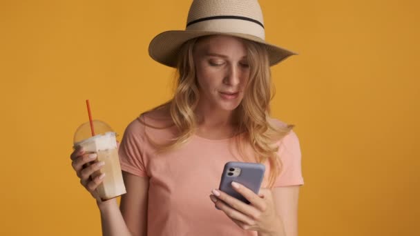 Csinos mosolygós szőke lány kalapban jeges kávét iszik boldogan használja okostelefon felett színes háttér
