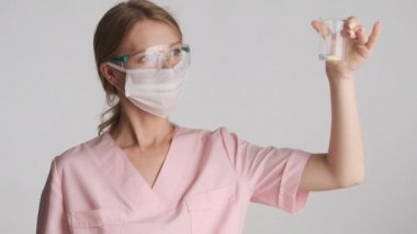 Koruyucu gözlüklü ve tıbbi maskeli genç bayan doktor gri arka planda yeni haplar üzerinde çalışıyor.