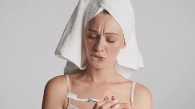 Başında havluyla genç ve çekici bir kadın. Düşünceli bir şekilde diş fırçasını izliyor ve gri arka planda mutlu bir şekilde gülümsüyor.