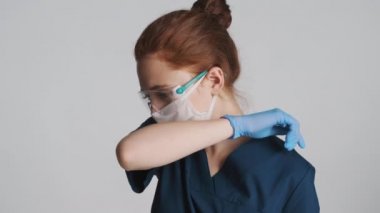 Koruyucu gözlüklü, maskeli ve eldivenli, omuzları gri arka planda öksüren kadın doktor.