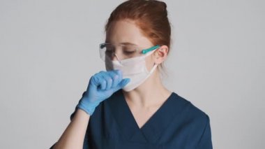 Koruyucu gözlüklü, maskeli ve beyaz arka planda öksüren tıbbi eldivenli kadın doktor. Kötü bir ifade.