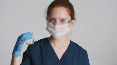 Koruyucu gözlüklü, maskeli ve tıbbi eldivenli güzel kadın doktor gri arka planda haplar gösteriyor.