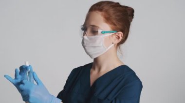 Koruyucu gözlüklü, maskeli ve tıbbi eldivenli kadın doktor beyaz arka planda kameraya antiseptik uyguluyor.