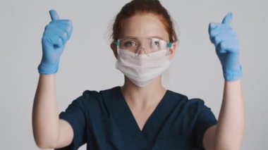 Koruyucu gözlüklü, maskeli ve tıbbi eldivenli çekici kadın doktor beyaz arka planda mutlu bir şekilde baş parmak hareketi gösteriyor.