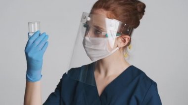 Koruyucu gözlüklü profesyonel kadın doktor, maske ve tıbbi eldivenler beyaz arka planda şişe tutuyor.