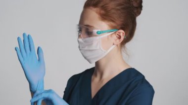 Koruyucu gözlüklü ve maskeli çekici, kendine güvenen kadın doktor beyaz arka planda tıbbi eldiven giyiyor.