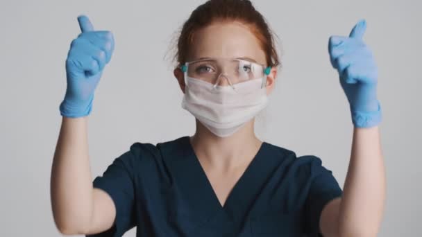 魅力的な女性医師で保護眼鏡 マスクと医療手袋喜んで上の親指を示すジェスチャー上の白い背景 — ストック動画