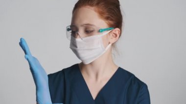 Koruyucu gözlüklü ve tıbbi maskeli genç, kendine güvenen bayan doktor gri arka planda tıbbi eldiven giyiyor.