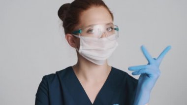 Koruyucu gözlüklü ve tıbbi maskeli genç çekici bayan doktor beyaz arka planda tıbbi eldiven giyiyor.