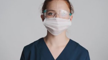 Koruyucu gözlüklü ve tıbbi maskeli genç bayan doktor gri arka plandan kameraya bakıyor. Güvenlik kavramı