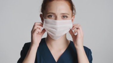 Genç, güzel bayan doktor maskesini çıkarıyor. Gri arka planda kameraya mutlu bir şekilde bakıyor. Güvenlik kavramı