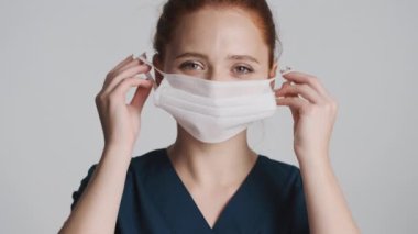 Tıbbi maske takan genç, güzel bir bayan doktor gri arka planda kameraya bakıyor. Güvenlik kavramı