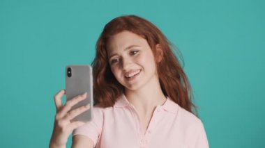 Güzel, gülümseyen, kızıl saçlı kız renkli arka planda akıllı telefondan arkadaşlarıyla görüntülü sohbet ediyor. Video çağrısı