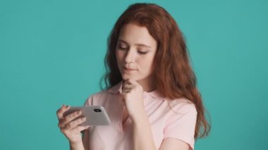 Çekici kızıl saçlı kız renkli arka planda akıllı telefondan video izliyor. Düşünme ifadesi