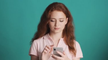 Güzel kızıl kız akıllı telefon kullanıyor ve renkli arka planda kameraya gülümsüyor.