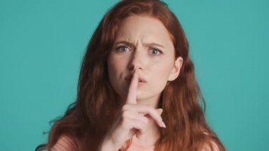 Çekici kızıl saçlı kız mavi arka planda kamerada sessizlik jesti yapıyor. İfade verme.