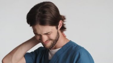 Genç, yorgun erkek doktor boyun ağrısı yüzünden masaj yapıyor ve beyaz arka planda kamerada gülümsedikten sonra...