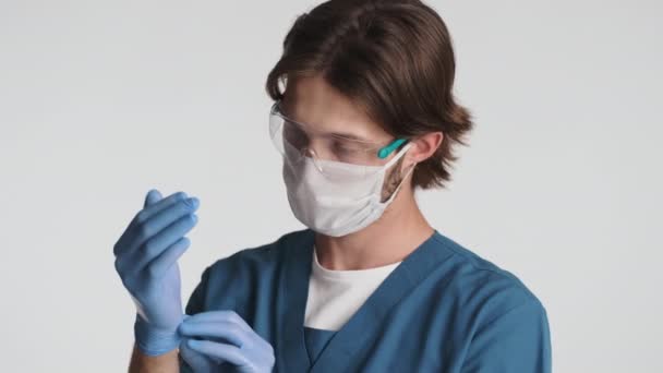 白い背景にカメラを楽しそうに見て手袋を身に着けている保護眼鏡や医療用マスクの若い魅力的な男性医師 安全コンセプト — ストック動画