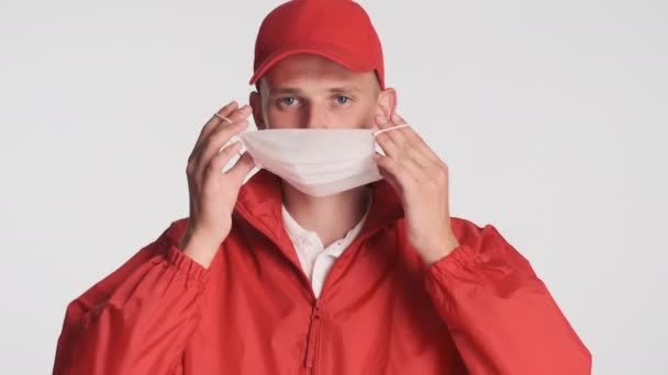 年轻的送货员头戴红帽子 身穿夹克衫 头戴医疗面罩 头戴白色背景 — 图库视频影像