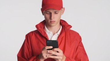Kırmızı şapkalı ve ceketli genç teslimatçının portresi beyaz arka planda akıllı telefon kullanıyor.